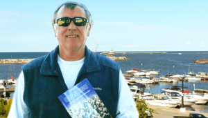 Mike McCrobie, with the Oswego lighthouse in the background, holds his second book — ‘We’re from Oswego ... and we couldn’t be any prouder!’ — which was published in November.